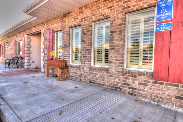 Exterior view of a brick building with four windows featuring white shutters and red wooden panels. There is a concrete walkway in front with a wooden planter box containing flowers and a yellow decorative flower. A bench is visible near the entrance, and a blue sign indicates reserved handicapped parking and van accessibility.