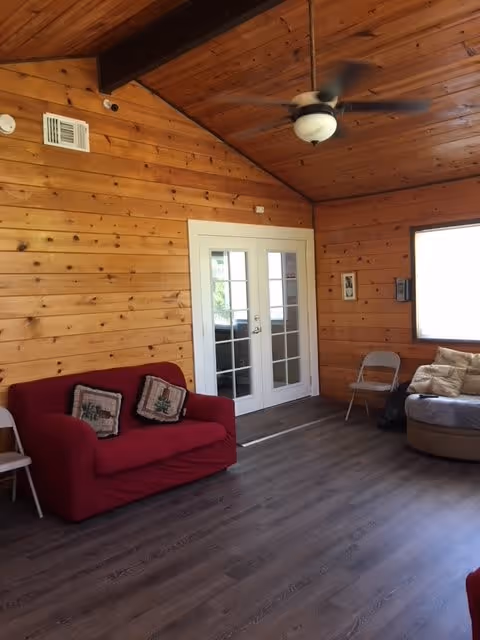 Interior of a room with wooden paneled walls and ceiling, featuring a ceiling fan, a red loveseat with two decorative pillows, a round cushioned chair with pillows, two folding chairs, and a set of white French doors.