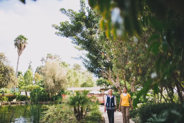 Two people walking on a shaded path beside a pond surrounded by trees and lush greenery.