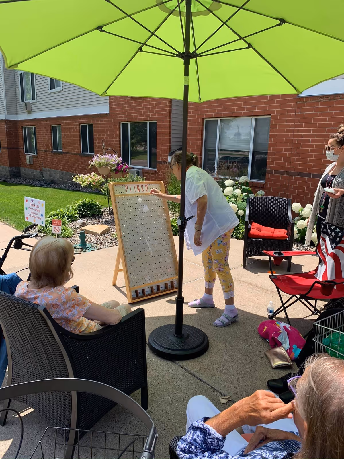 Several elderly women are gathered outside under a large green umbrella playing a game of Plinko on a wooden board. One woman is standing and dropping a disc onto the board, while others are seated around watching. The setting is a patio area next to a brick building with windows and some landscaping with flowers and greenery.