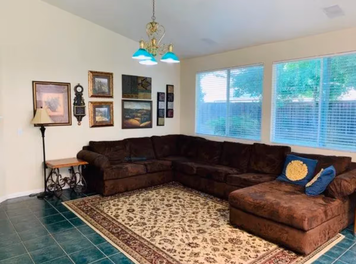 A cozy living room with a large brown sectional sofa adorned with blue and yellow pillows. The floor is covered with a patterned area rug, and the walls are decorated with framed pictures and a wall clock. There is a floor lamp and a small side table next to the sofa. Large windows with blinds allow natural light to brighten the room.