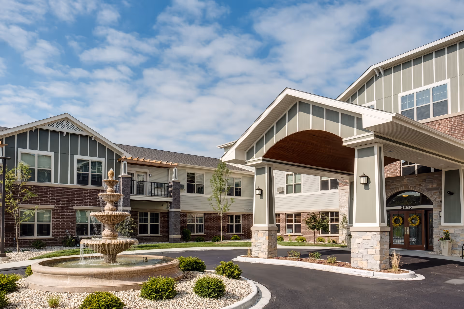 Exterior view of Heritage Lake Country Senior Living facility showing a large covered entrance with stone pillars, a multi-tiered water fountain in front, and a two-story building with brick and siding under a partly cloudy sky.