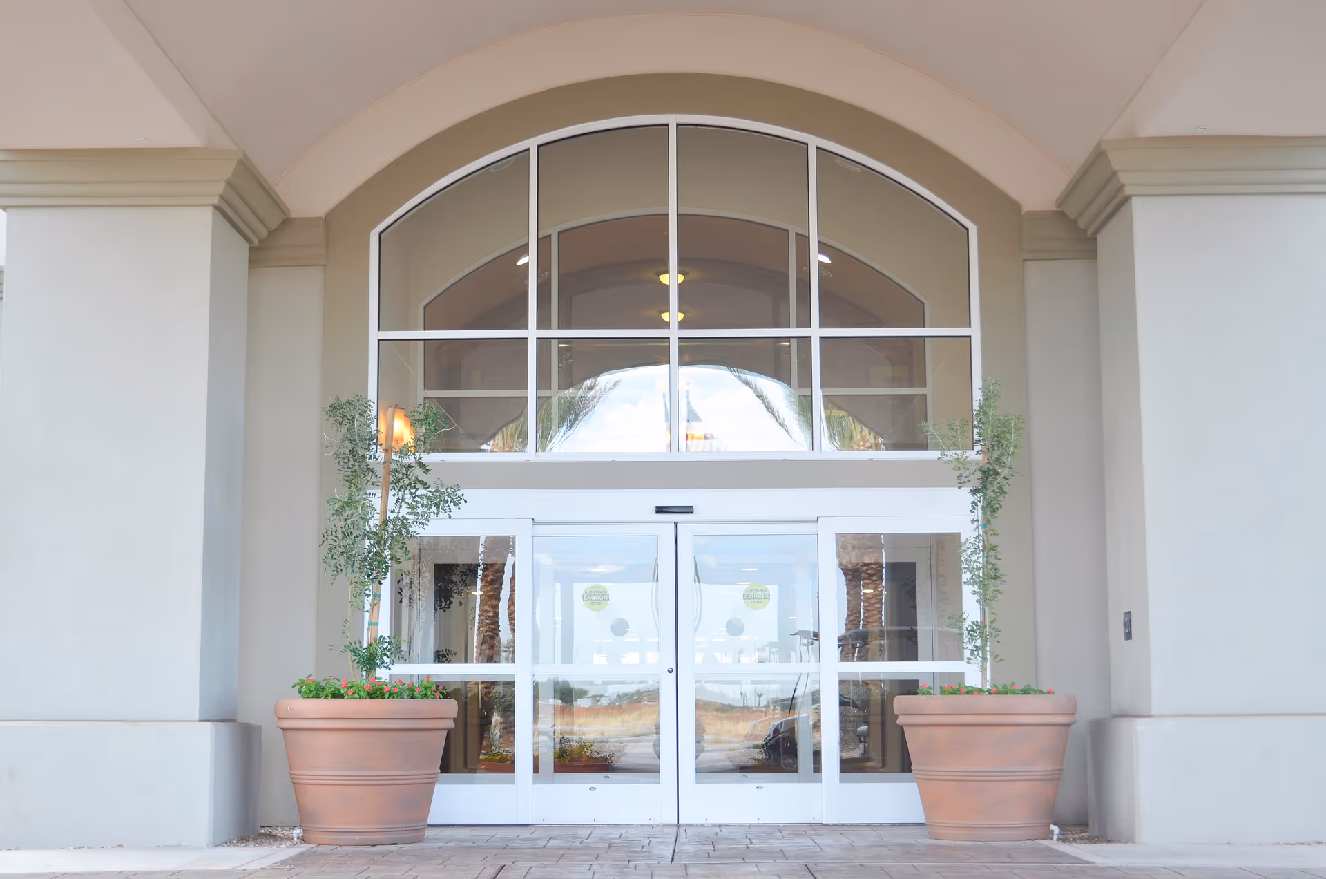 Entrance of a building with large glass double doors framed by an arched window above. Two large terracotta planters with small trees flank the doorway. The exterior walls are light-colored with decorative molding.