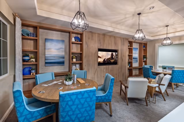 A cozy and modern common area in a senior living facility featuring a round wooden table with blue upholstered chairs around it, playing cards and a game on the table, a wall-mounted TV, built-in wooden shelves with decorative items, and additional seating with white and blue chairs near a window.