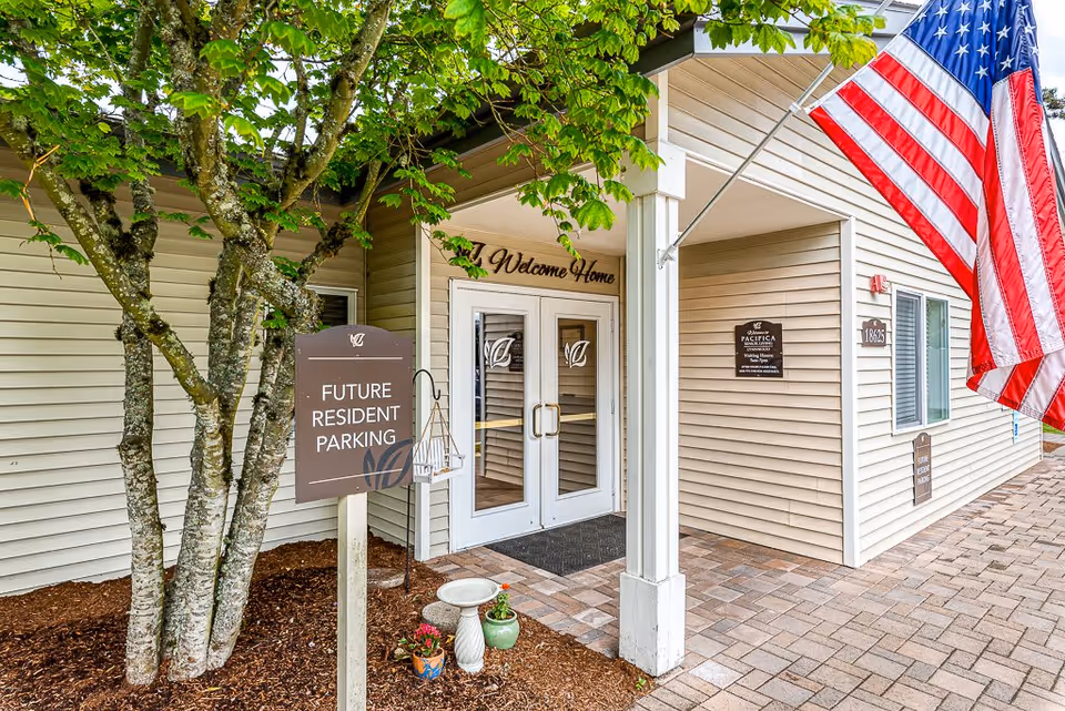 Entrance to a senior living facility with double glass doors under a covered porch. A sign near the entrance reads 'Future Resident Parking.' There is a large American flag mounted on the right side of the building. Trees and small potted plants are visible near the entrance, and a 'Welcome Home' sign is above the doors.