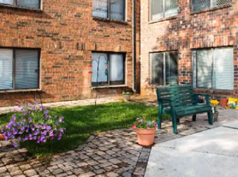 Outdoor courtyard area with brick building walls, windows with blinds, a green bench, potted plants, purple flowers, and a bird feeder.