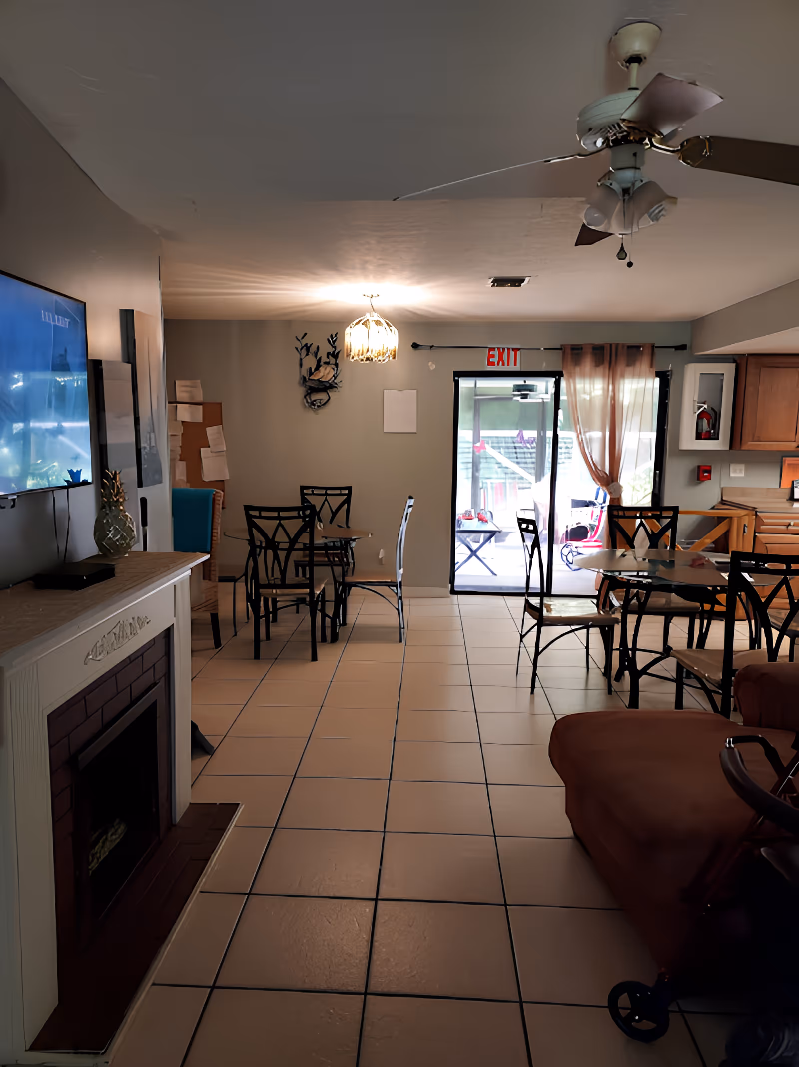 Interior view of a common area in a senior living facility with tiled floors, several glass-top tables with metal chairs, a ceiling fan, a wall-mounted TV above a decorative fireplace, and a sliding glass door leading to an outdoor patio.