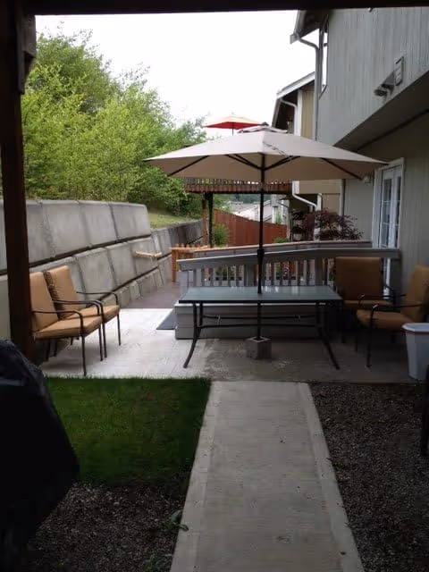 Outdoor patio area with a table and umbrella, surrounded by cushioned chairs. The patio is adjacent to a building with a concrete walkway leading to the seating area. There is greenery and trees in the background.