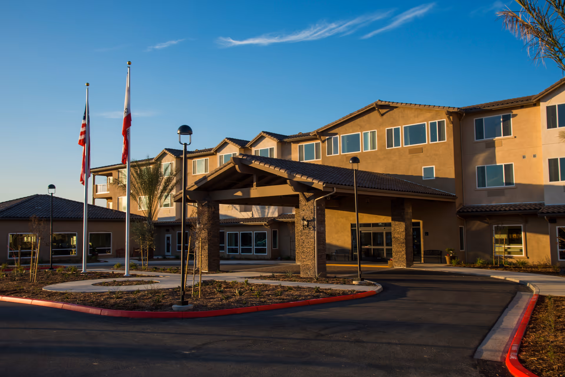Exterior view of Renaissance Village Murrieta senior living facility showing the main entrance with a covered driveway, flagpoles with flags, and a clear blue sky.