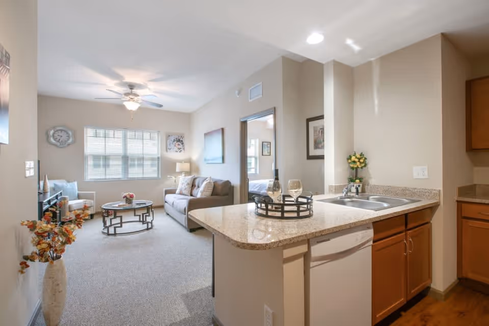 A bright and cozy living area with a beige sofa, a round coffee table, and a window with blinds. The room has light-colored walls and carpeted floor. To the right is a kitchen area with a granite countertop, a sink, wooden cabinets, and a dishwasher. A bedroom is visible through an open doorway.