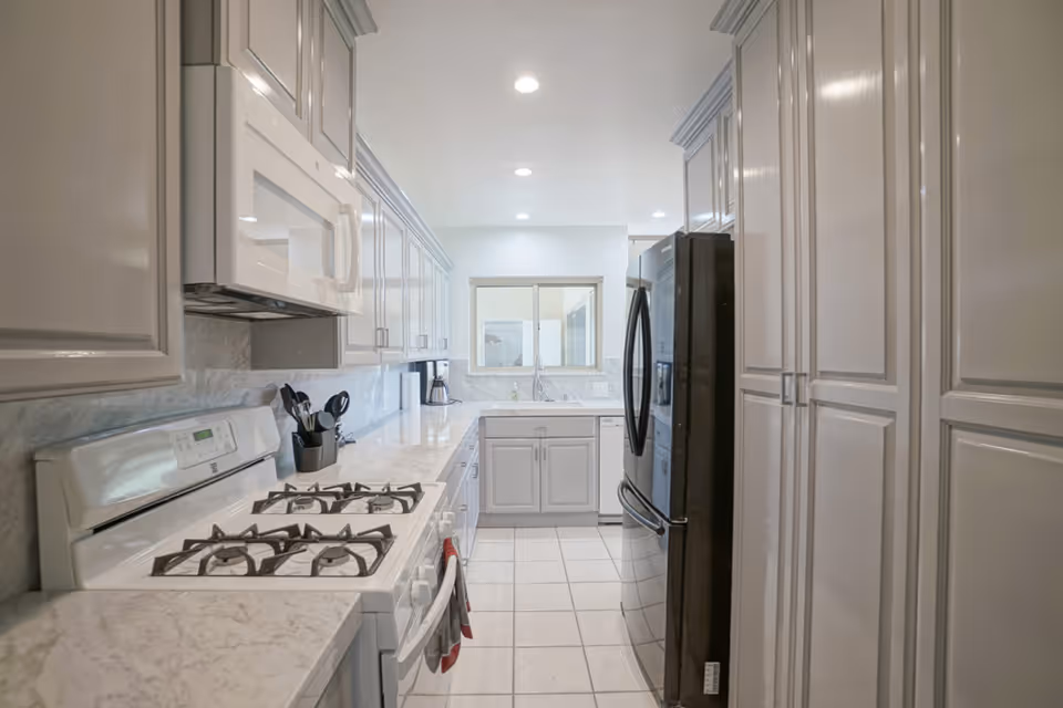 A bright, modern kitchen with white cabinetry, marble countertops, a white gas stove with four burners, a white microwave above the stove, a black refrigerator, and a coffee maker on the counter. The kitchen has tiled flooring and recessed ceiling lights.