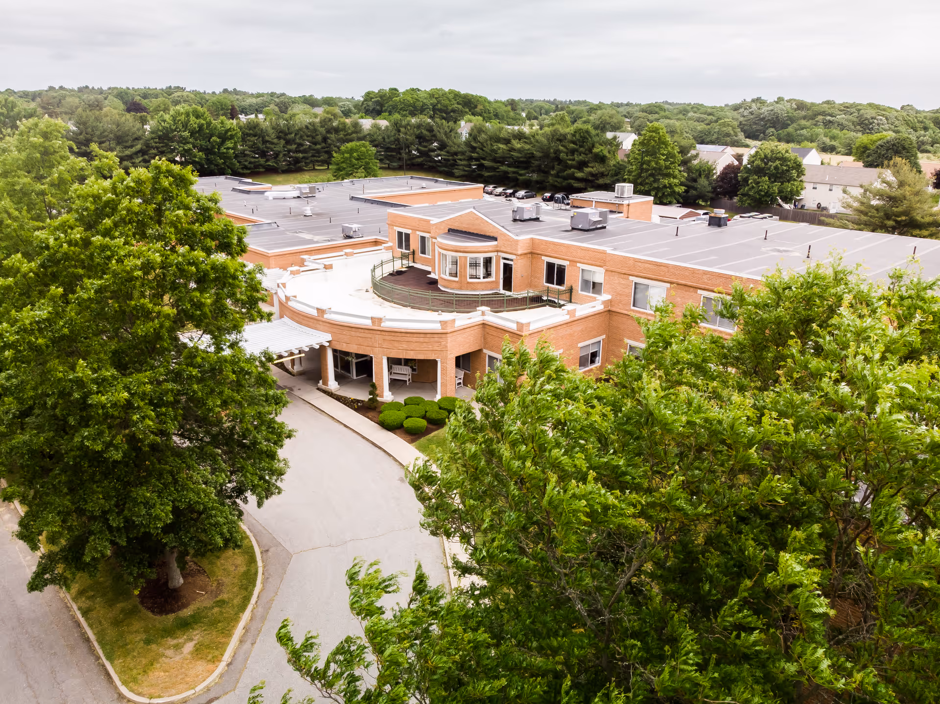 Aerial view of CareOne at New Bedford, a large brick building surrounded by trees and greenery, with a driveway leading to the entrance under a covered portico.