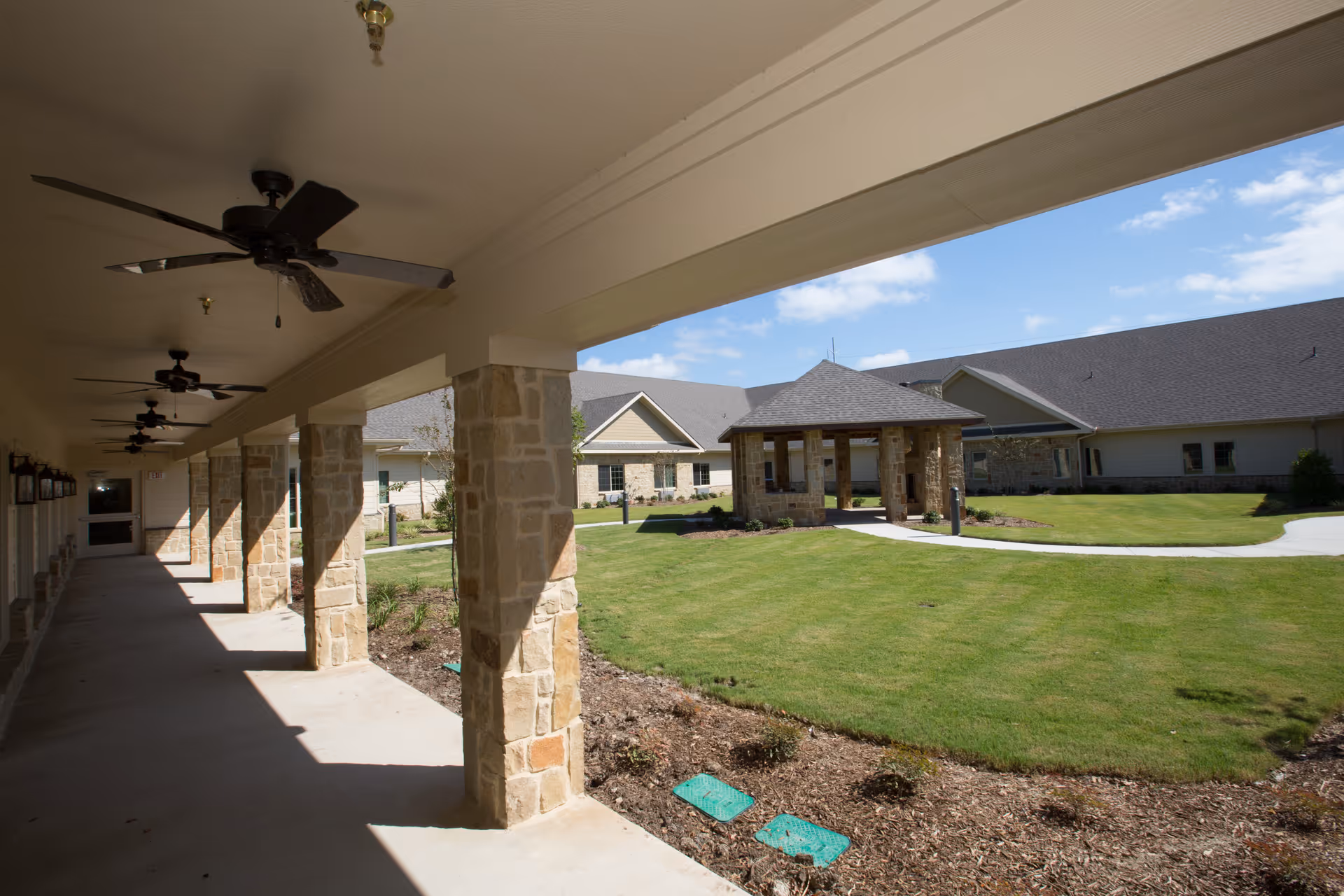 Covered outdoor walkway with stone pillars and ceiling fans overlooking a green lawn and a gazebo structure, with a building in the background under a blue sky with some clouds.