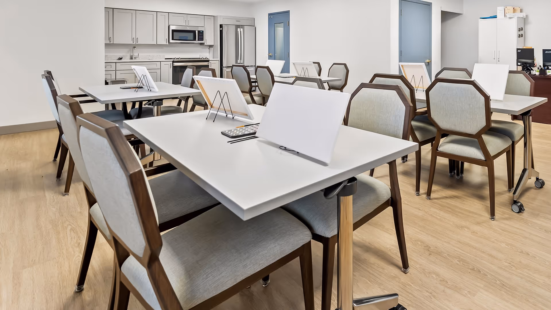 A bright and clean dining area in a senior living facility with several white tables and cushioned chairs arranged neatly. Each table has a white display stand and a remote control. In the background, there is a modern kitchen area with white cabinets, a microwave, and a refrigerator. The room has light wood flooring and white walls.
