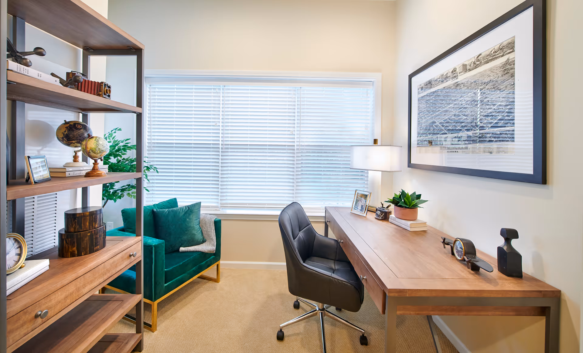 A bright office or study room with a large window covered by white blinds. The room features a wooden desk with a black swivel chair, a table lamp, framed pictures, a small plant, and decorative items. To the left, there is a green upholstered armchair with a throw blanket and a wooden shelving unit holding globes, books, and other decor.