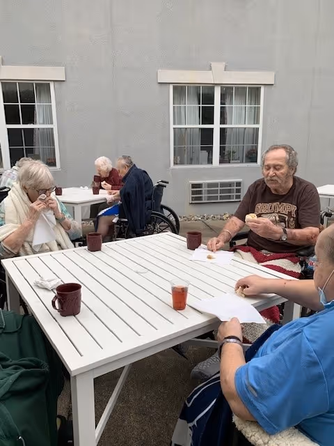 Four elderly individuals sitting around a white outdoor table with drinks and snacks, two of them in wheelchairs, outside a building with gray walls and windows.