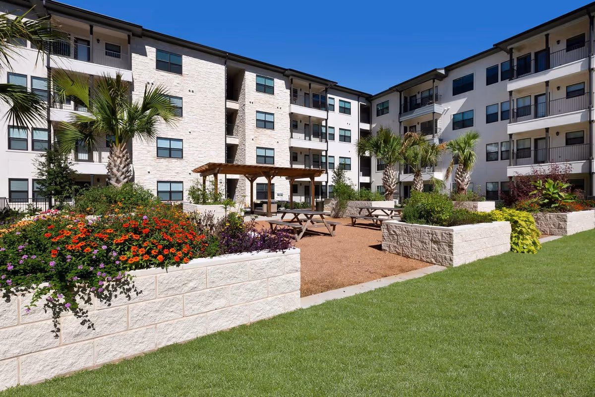 Outdoor courtyard area of a senior living facility with raised flower beds filled with colorful flowers and greenery, several palm trees, picnic tables, and a wooden pergola. The courtyard is surrounded by a multi-story building with balconies and windows under a clear blue sky.