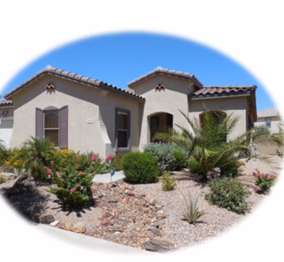 Exterior view of a single-story beige stucco building with a tiled roof, surrounded by desert landscaping including various shrubs, small palm trees, and flowering plants under a clear blue sky.