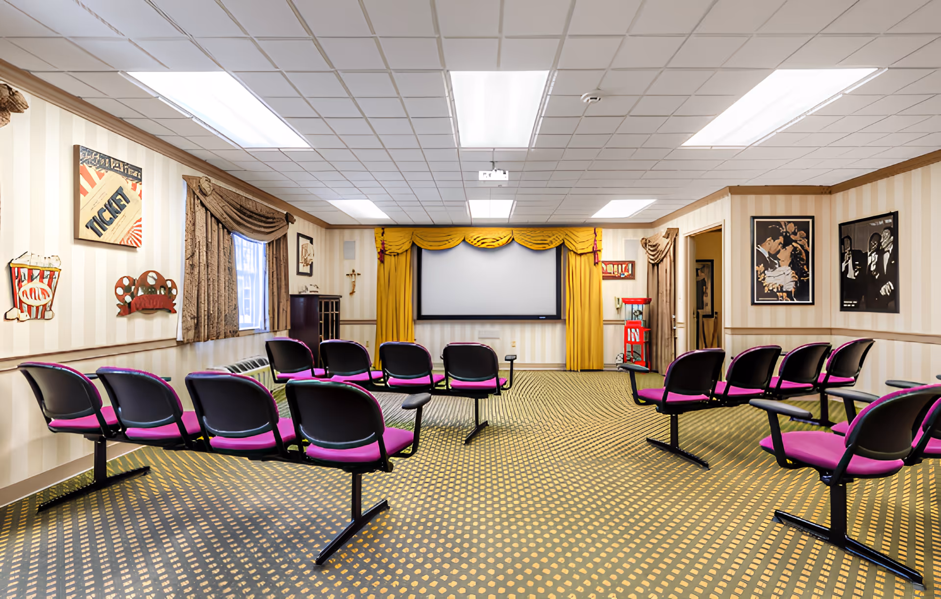 A senior living facility's small theater room with rows of purple cushioned chairs facing a large screen framed by yellow curtains. The room has patterned carpet, beige striped wallpaper, framed movie-themed artwork, and a popcorn machine in the corner.
