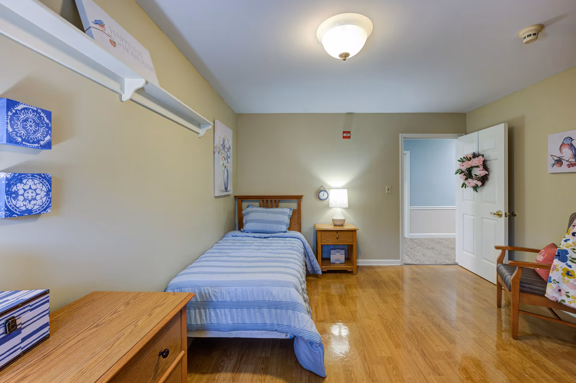 A simple bedroom with a single bed covered in blue striped bedding, a wooden nightstand with a lamp, a wooden dresser, and a chair with a floral cushion. The walls are beige with some artwork, and the door is open showing a hallway with light blue walls.
