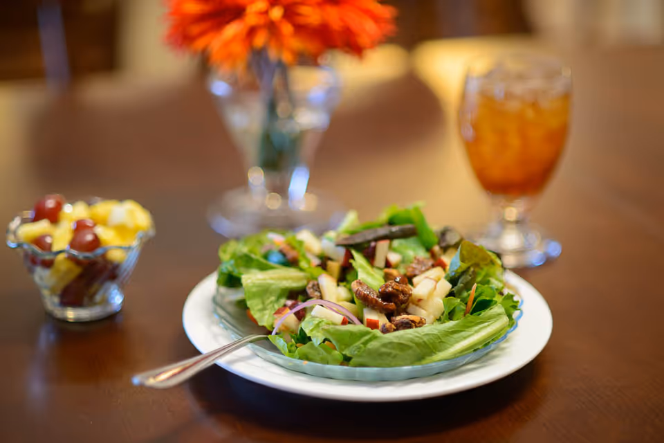 A close-up of a fresh salad with lettuce, apple slices, pecans, and red onions on a white plate with a fork. In the background, there is a glass bowl with fruit salad, a glass of iced tea, and a vase with orange flowers on a wooden table.