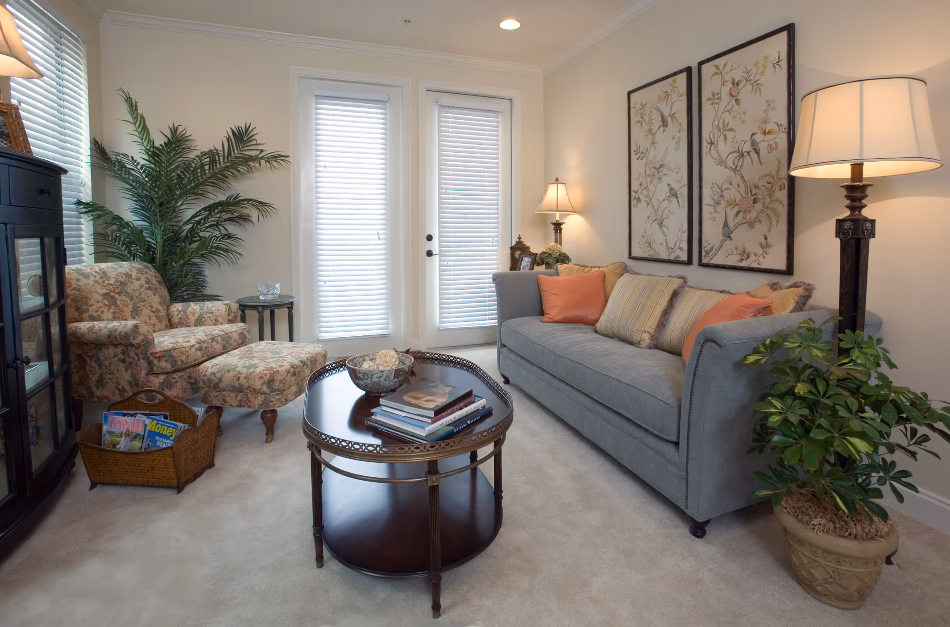 A cozy living room with a gray sofa adorned with multiple cushions, a floral armchair with matching ottoman, a round wooden coffee table with books and a decorative bowl, two framed floral artworks on the wall, a tall floor lamp, a potted plant, and a basket with magazines. French doors with blinds allow natural light into the room.
