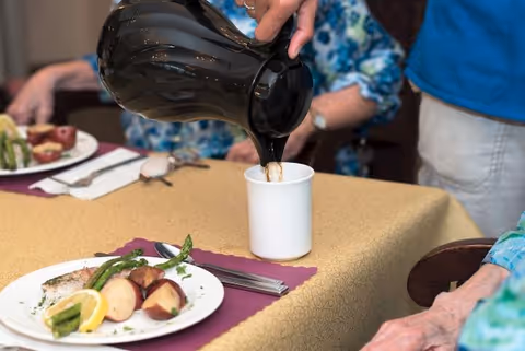 A person pours coffee from a black carafe into a white mug at a dining table set with plates of food.