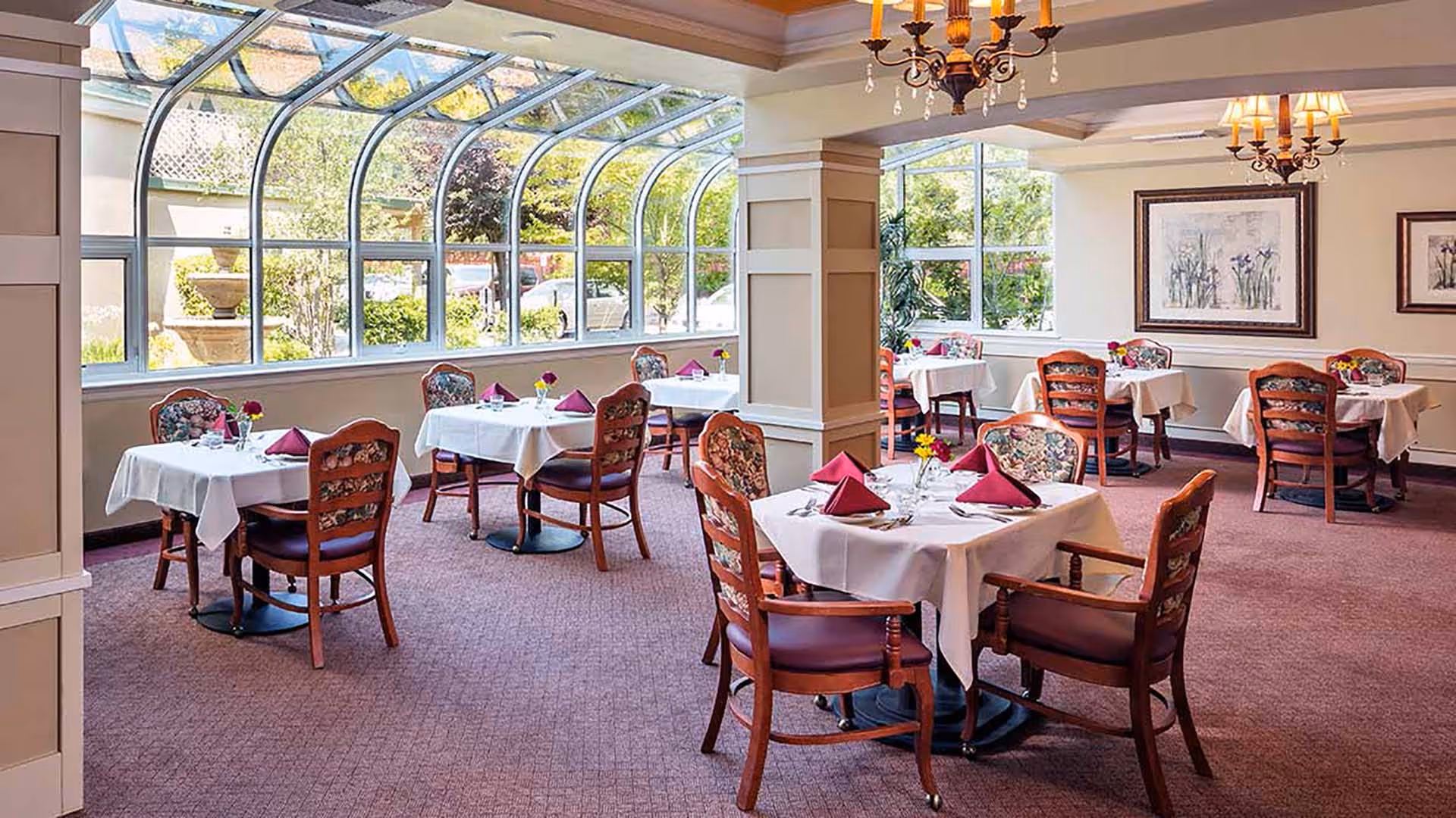 A bright dining room with several tables covered in white tablecloths, each set with folded burgundy napkins, glassware, and small flower arrangements. The room features large windows and a glass ceiling section allowing natural light to fill the space. Wooden chairs with floral upholstery surround the tables, and chandeliers hang from the ceiling.