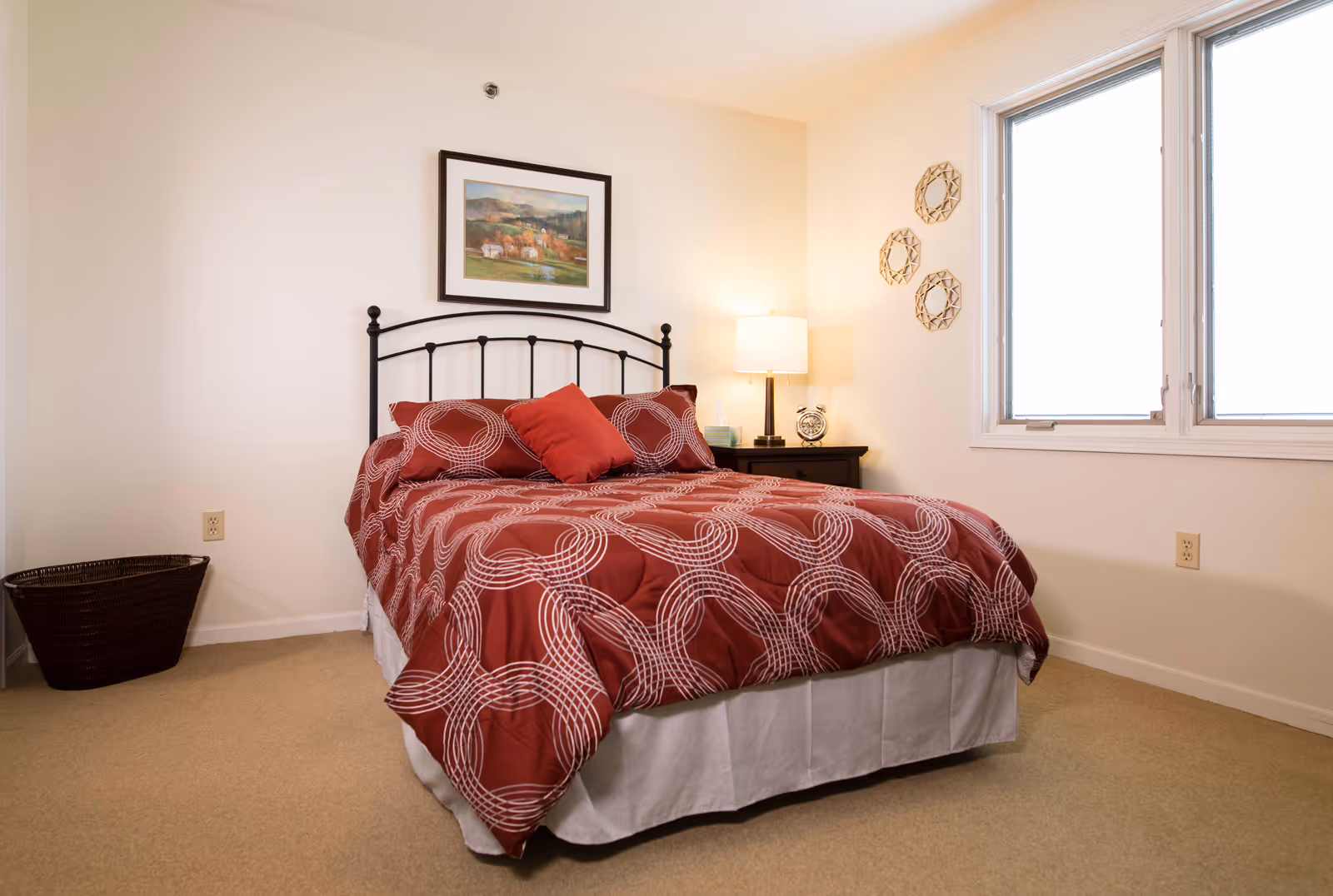 A bedroom with a bed featuring a red and white patterned comforter and pillows. The bed has a black metal headboard. There is a nightstand with a lamp and an alarm clock next to the bed. A framed landscape painting hangs on the wall above the bed. Three decorative circular wall hangings are on the adjacent wall near a window with white blinds. A dark woven basket is placed on the floor in the corner.