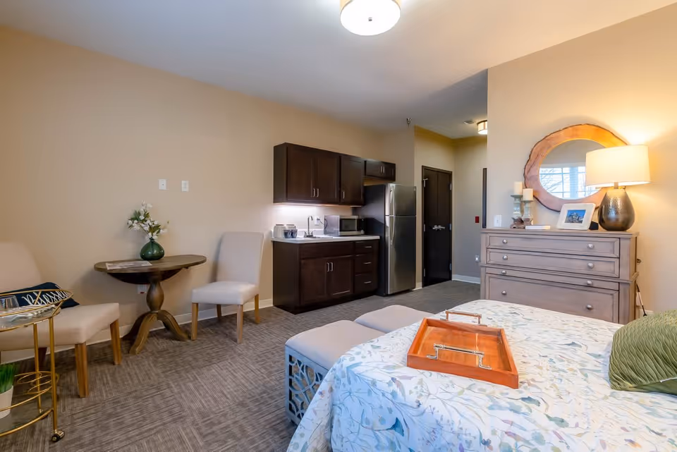 A cozy room with a bed covered in a floral bedspread and a green pillow, a wooden tray on the bed, two cushioned stools at the foot of the bed, a wooden dresser with a round mirror above it, a table lamp, and decorative items. To the left, there is a small round table with two chairs and a vase with flowers. The room also features a kitchenette with dark wood cabinets, a microwave, a toaster, and a stainless steel refrigerator. The walls are painted beige and the floor is carpeted.