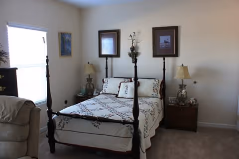 A bedroom with a four-poster bed covered with a white quilt featuring a floral pattern. There are two matching bedside tables with lamps on each side of the bed. The walls are light-colored and decorated with three framed pictures. A window with blinds is on the left side, and part of a beige armchair is visible in the foreground.