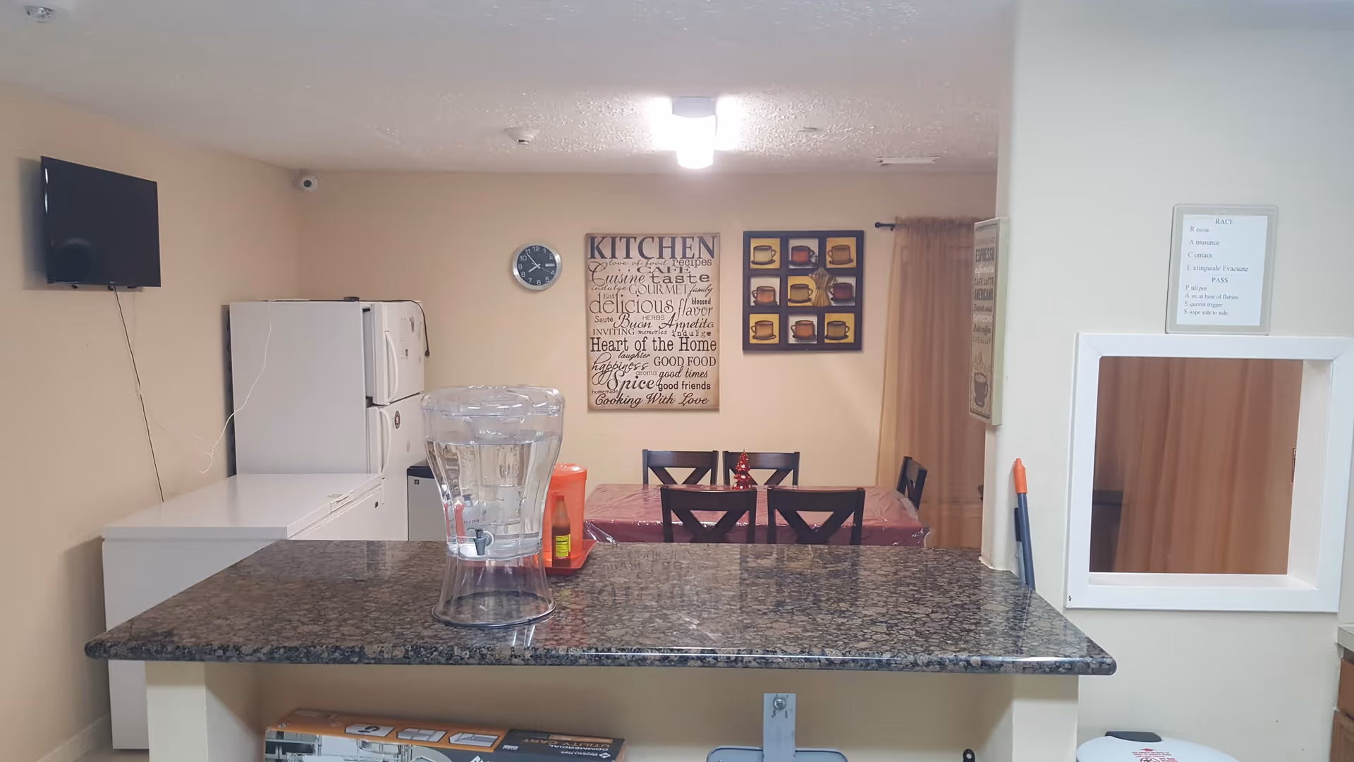 Interior view of a kitchen and dining area in an assisted living facility. The foreground shows a granite countertop with a clear water dispenser and a bottle of ketchup. In the background, there is a dining table with six chairs, a wall clock, and two framed wall decorations. On the left side, there is a white refrigerator and freezer, and a wall-mounted TV. The walls are beige, and there is a small window with a curtain on the right.