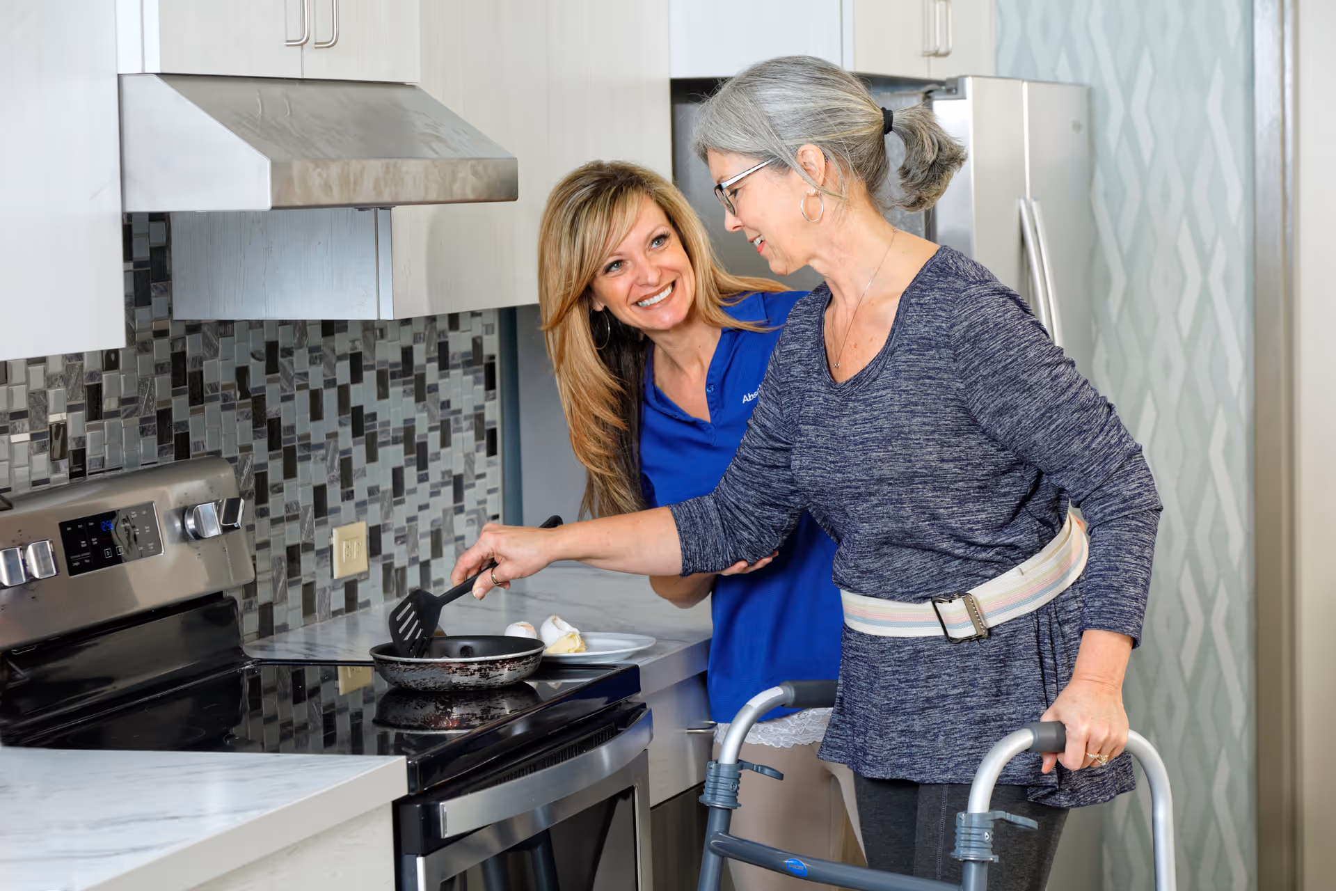 A caregiver assists an elderly woman using a walker as she cooks at a stove in a modern kitchen.