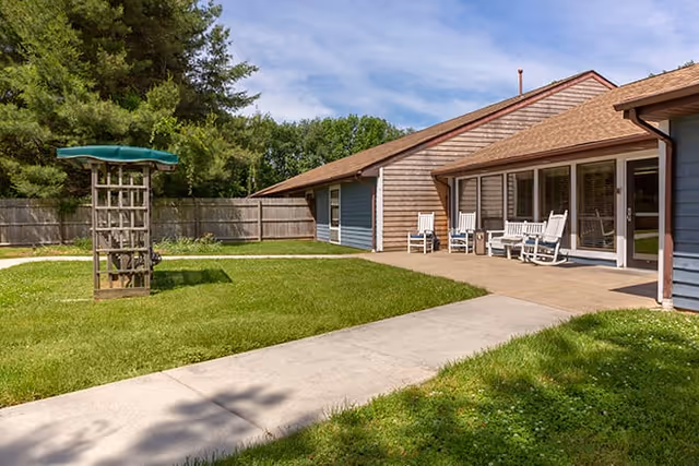 Outdoor patio area of a senior living facility with a concrete walkway, green grass, wooden fence, and a small wooden structure with a green canopy. The building has a sloped roof and a covered porch with white rocking chairs.