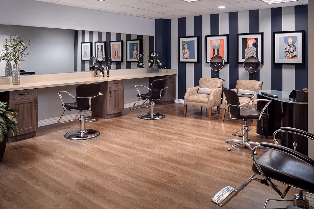 Interior of a salon area with wooden flooring, three black salon chairs, two beige armchairs, and a long counter with mirrors and hair dryers mounted on the wall. The walls have vertical black and white stripes and framed artwork featuring stylized portraits.