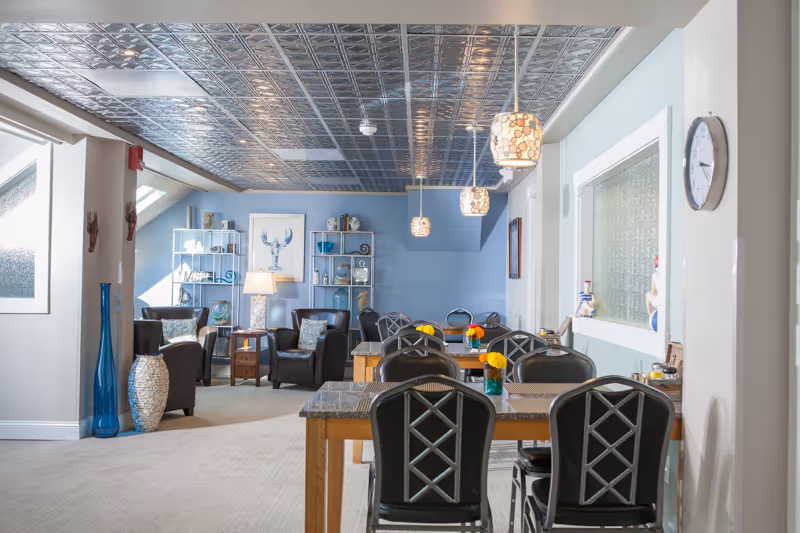 Bright communal dining and sitting area with tables and chairs in the foreground, leather armchairs and blue shelving in the background, and decorative pendant lights overhead.
