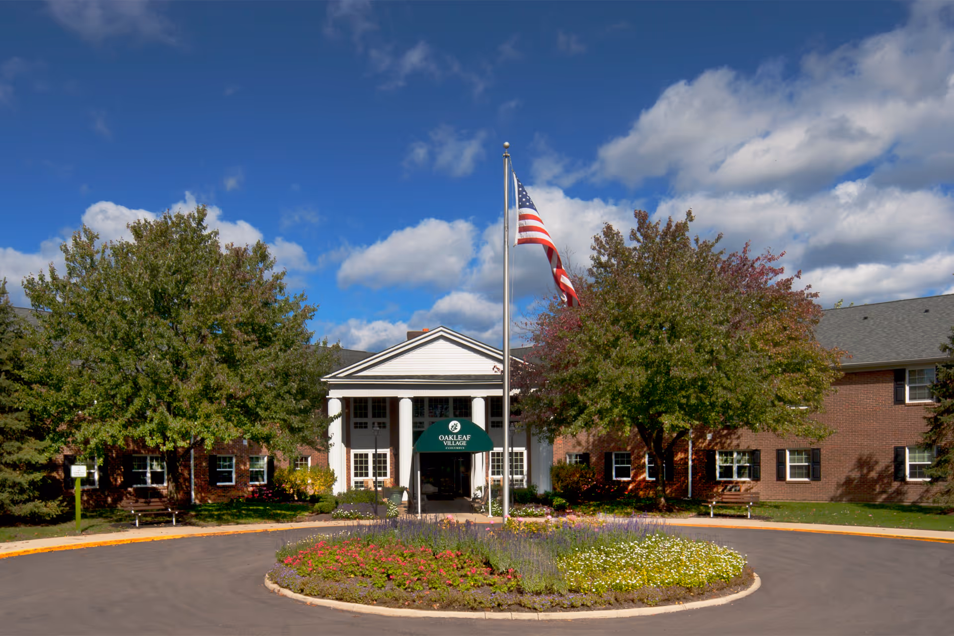 Front entrance of a brick building with white columns, an American flag, a circular driveway and a landscaped flower bed under a blue sky.