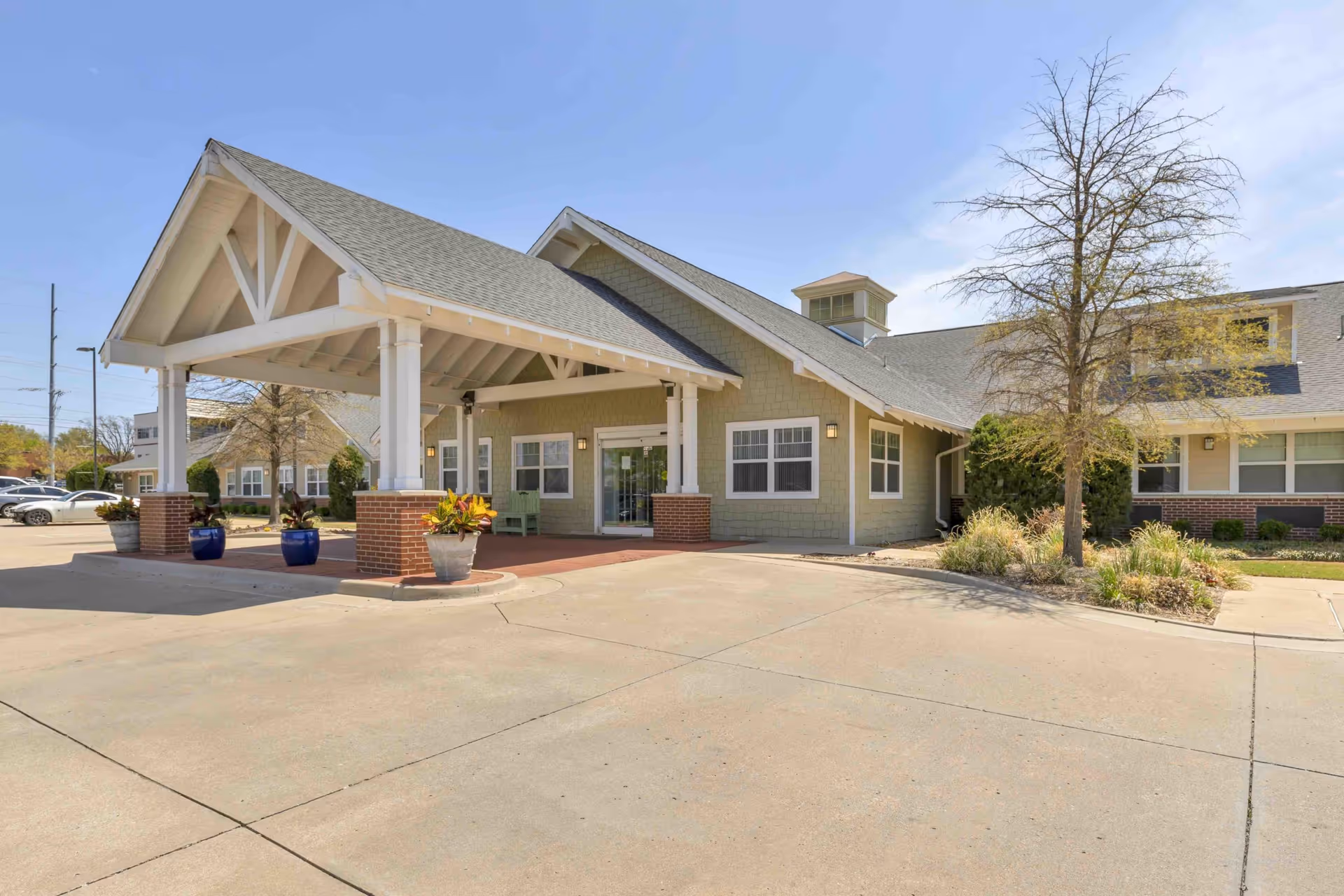 Exterior view of Aberdeen Memory Care of Tulsa building entrance with a covered drop-off area supported by white columns with brick bases, potted plants, a tree, and a clear blue sky.