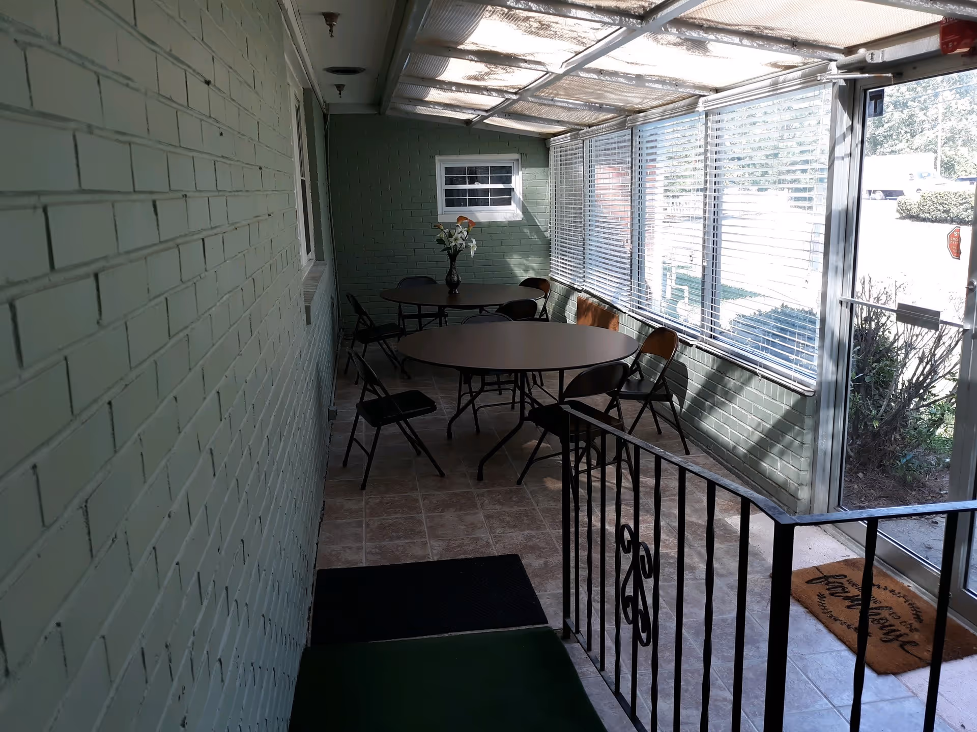 A sunroom or enclosed porch area with green painted brick walls, tiled floor, and a glass door leading outside. The room contains two round tables with several folding chairs around them, and a vase with flowers on one table. Large windows with blinds allow natural light to enter the space.