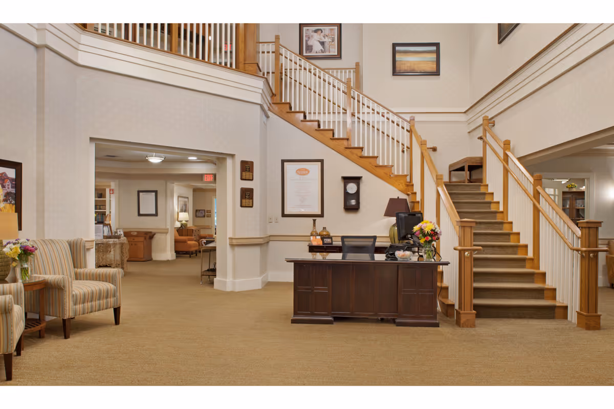 Interior view of a senior living facility lobby featuring a wooden reception desk with a computer and flower vase, a staircase with wooden handrails and carpeted steps leading to an upper floor, two striped armchairs with a small side table holding a flower vase, and framed artwork on the walls.