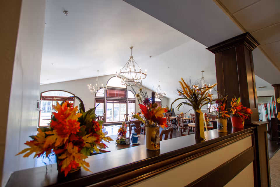 Interior view of a dining room in a senior living facility with multiple wooden tables and chairs, decorated with several vases of colorful autumn-themed flowers on a wooden partition. Large windows with arched tops allow natural light to fill the room, and chandeliers hang from the ceiling.