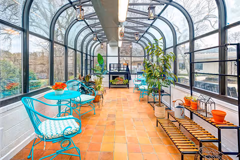 A bright sunroom with large arched windows and a glass ceiling, featuring turquoise metal chairs with patterned cushions, small round tables, various potted plants, and a wooden bench with gardening items on a terracotta tiled floor.