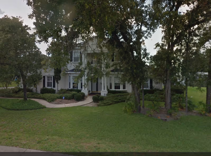 Two-story white senior living building with columns, landscaped yard, and trees.