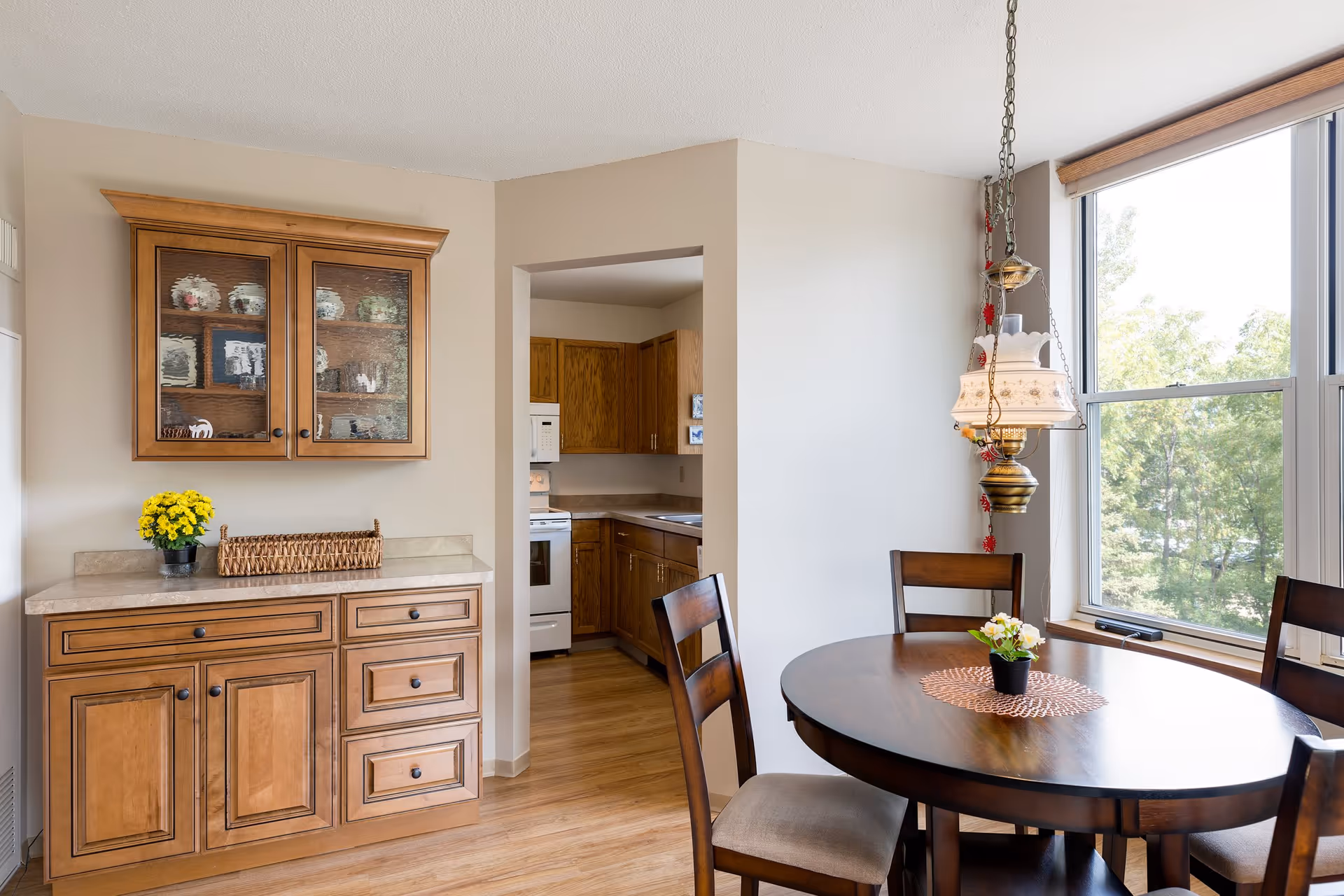 A bright dining area with a round wooden table and four chairs, a small potted plant on the table, a hanging decorative lamp, a large window showing green trees outside, and a wooden cabinet with glass doors displaying decorative items. The dining area opens into a kitchen with wooden cabinets and white appliances.