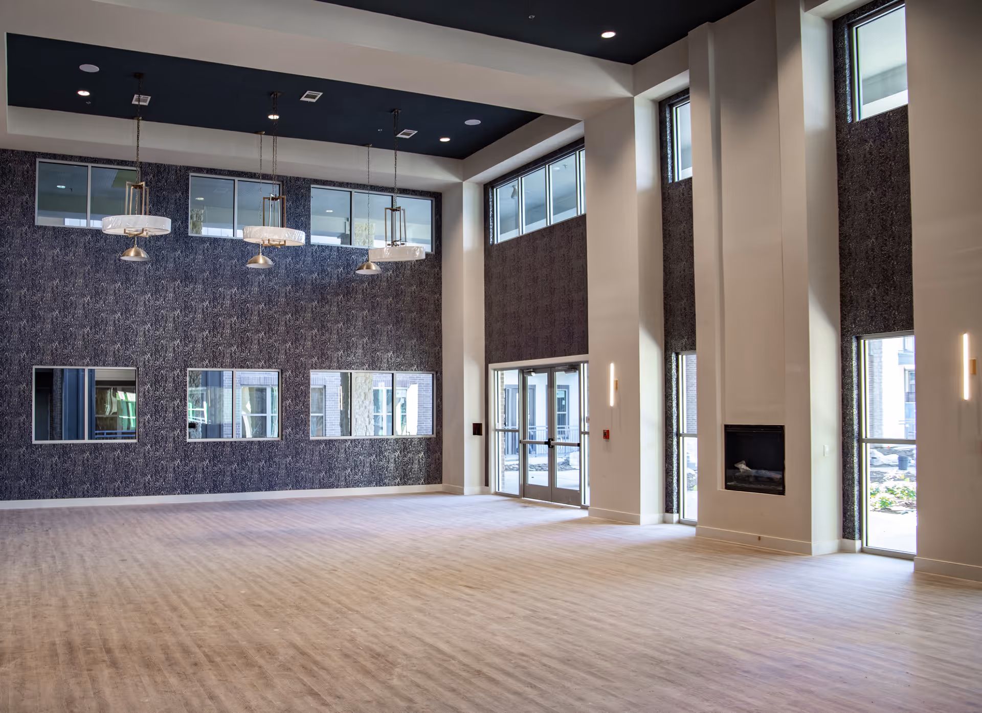 Spacious, modern interior room with high ceilings, large windows, and a patterned dark accent wall. The room features hanging pendant lights, a fireplace, and glass double doors leading outside. The floor is light-colored wood, and the space is empty.
