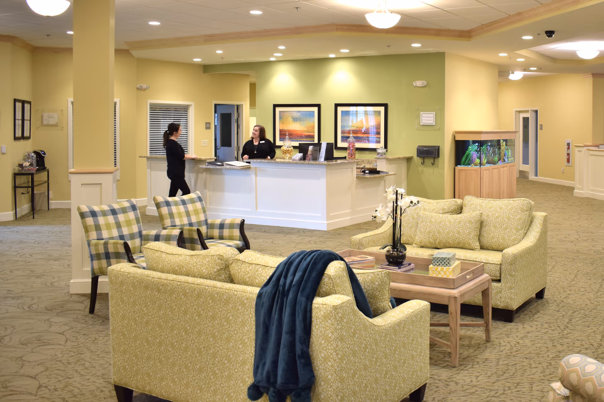 A bright and welcoming senior living facility lobby with a seating area consisting of two patterned armchairs and two green sofas around a wooden coffee table. A blue throw blanket is draped over one sofa. In the background, two staff members are conversing behind a white reception desk with decorative items and framed artwork on the wall. To the right, there is a large fish tank and hallways leading to other rooms.