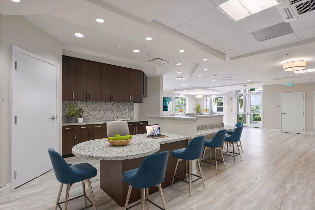 Bright communal kitchen and seating area with granite countertops, blue bar stools, and dark wood cabinets in a senior living facility.