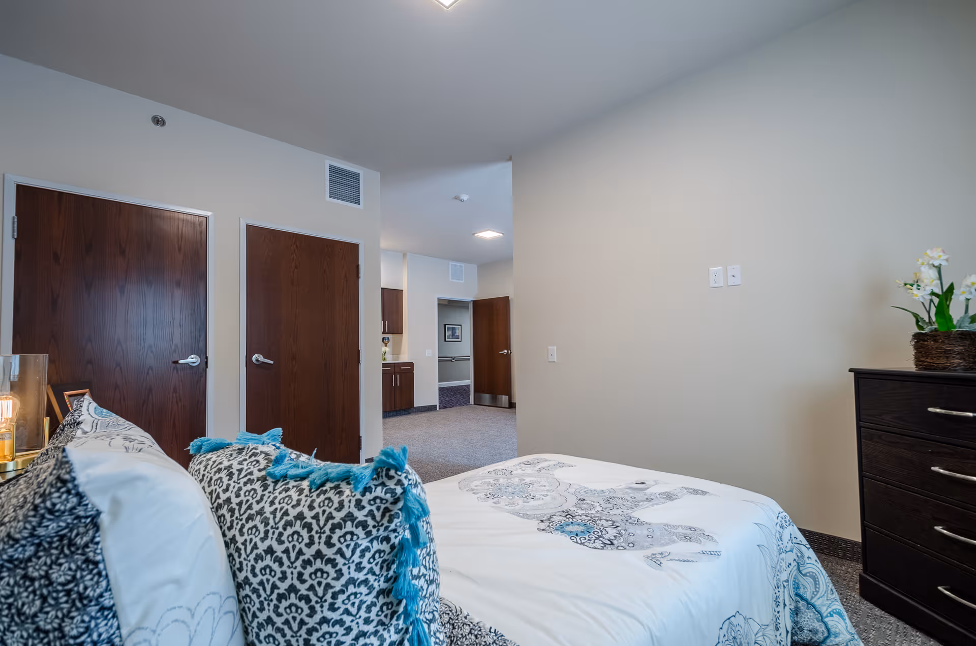 A bedroom in a senior living facility featuring a bed with patterned pillows and a white and blue bedspread. There are two closed wooden doors on the left wall, a dark wooden dresser with a plant on top on the right, and an open doorway leading to a hallway with cabinets and another door in the background.