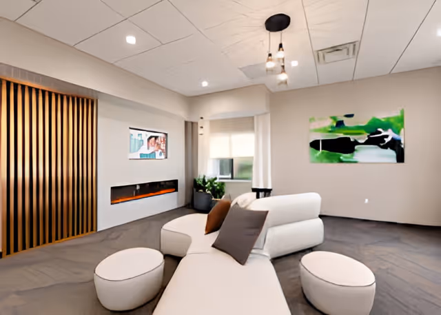 Modern lounge with a curved white sofa and ottomans facing a wall-mounted TV and electric fireplace, with a green abstract painting on the wall.