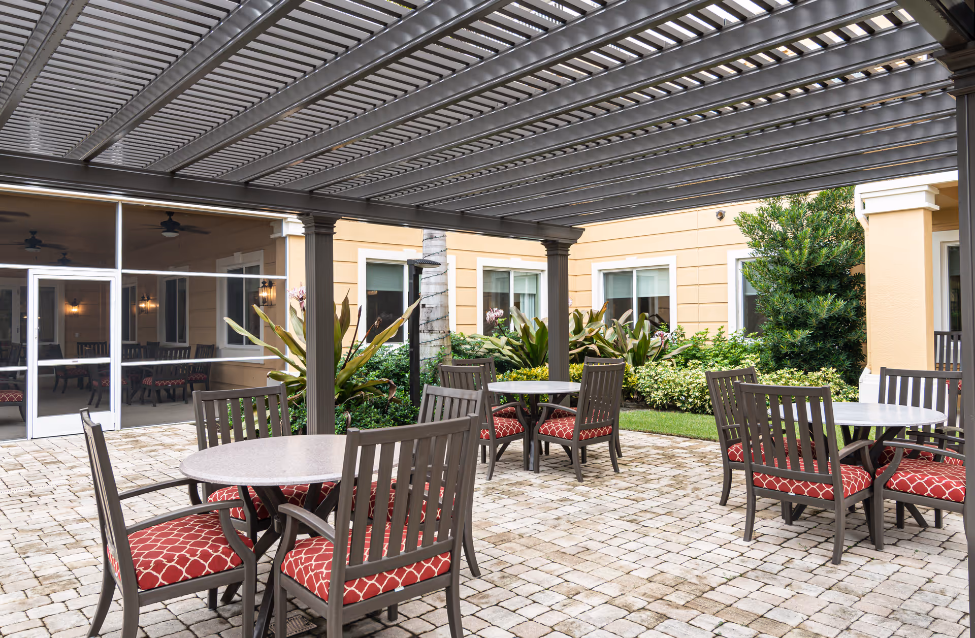 Outdoor patio area with round tables and chairs featuring red patterned cushions, under a metal pergola. The patio is paved with stone tiles and surrounded by plants and shrubs, with a building featuring large windows in the background.